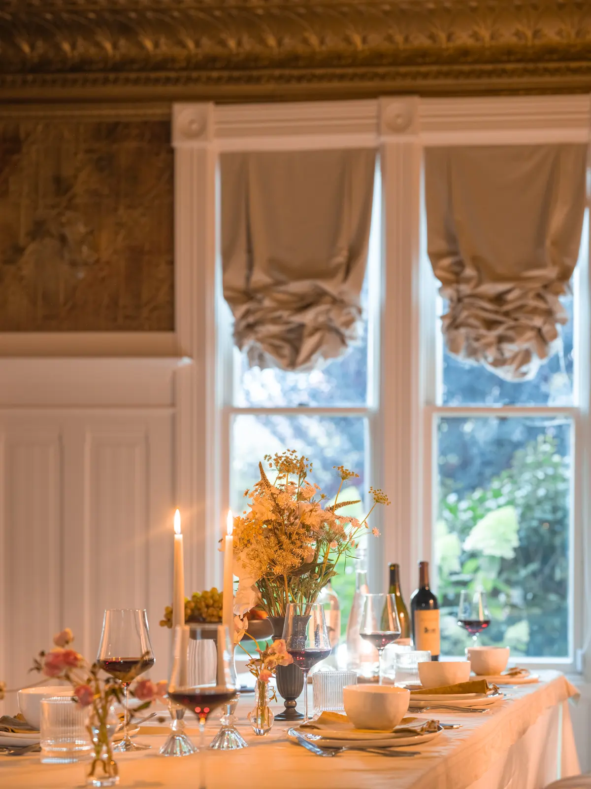 Dining table with candles and flowers beside tall windows.