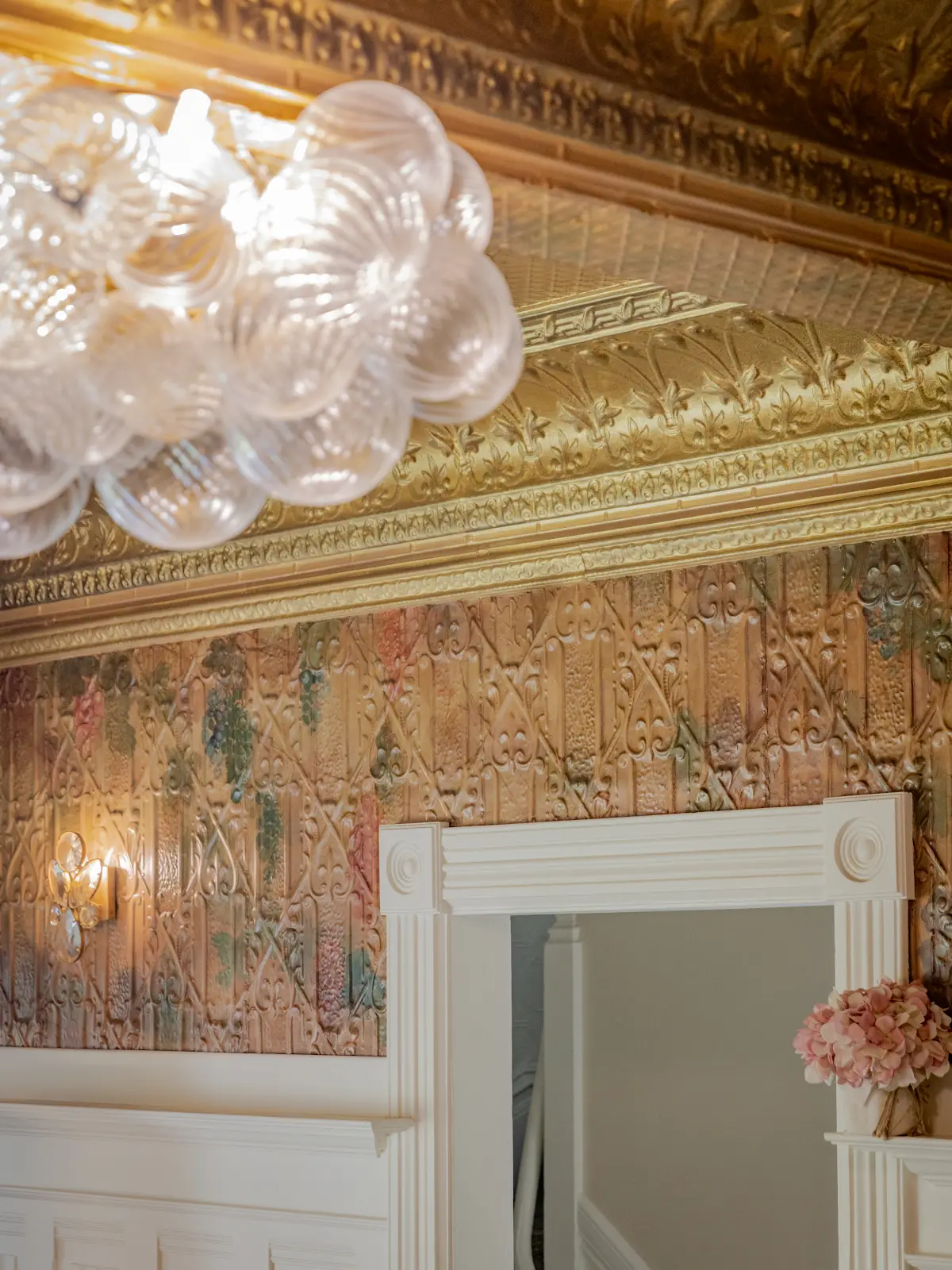 Ornate gold ceiling, glass chandelier, and patterned wallpaper above a doorway.